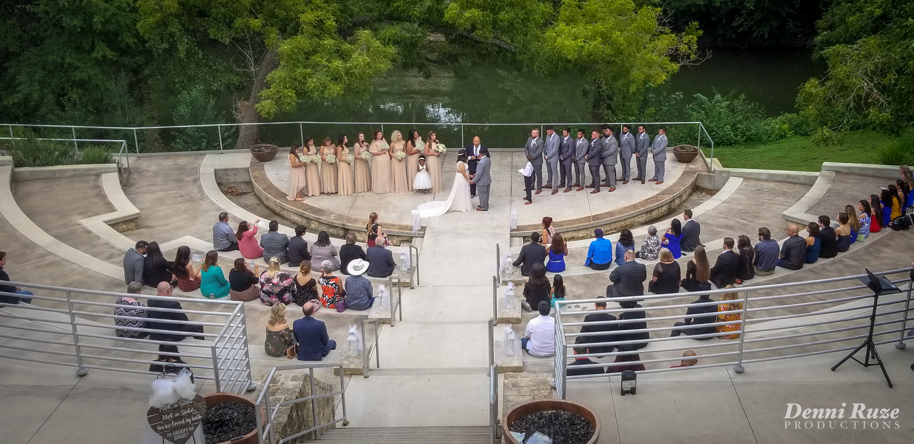 The wedding ceremony, with the bridal party on stage and the bride and groom listening to the minister. Guests are seated, with green foliage and river in the background.